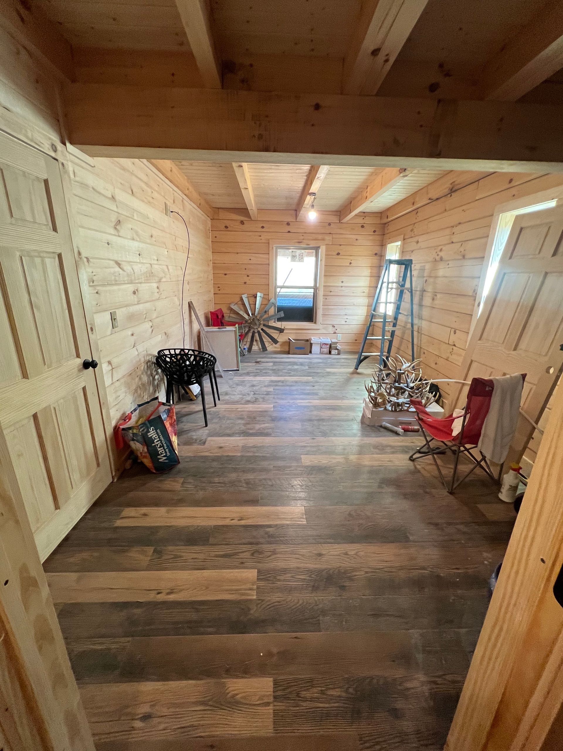 Rustic unfinished room with wood walls and floor, scattered chairs, ladder, and tools near a window.