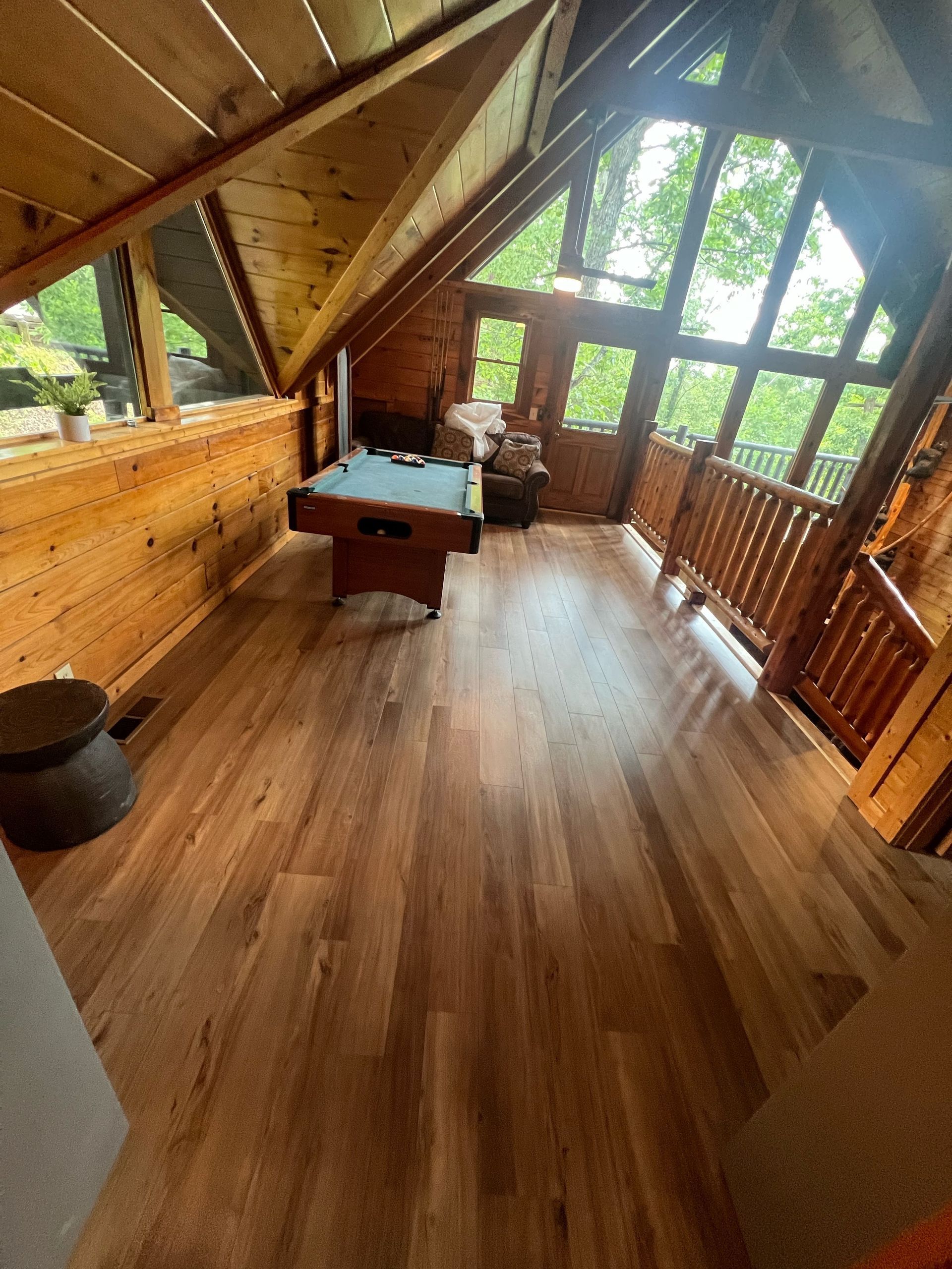 Wood-paneled attic room with a sloped ceiling, wooden floor, desk, and open windows overlooking greenery
