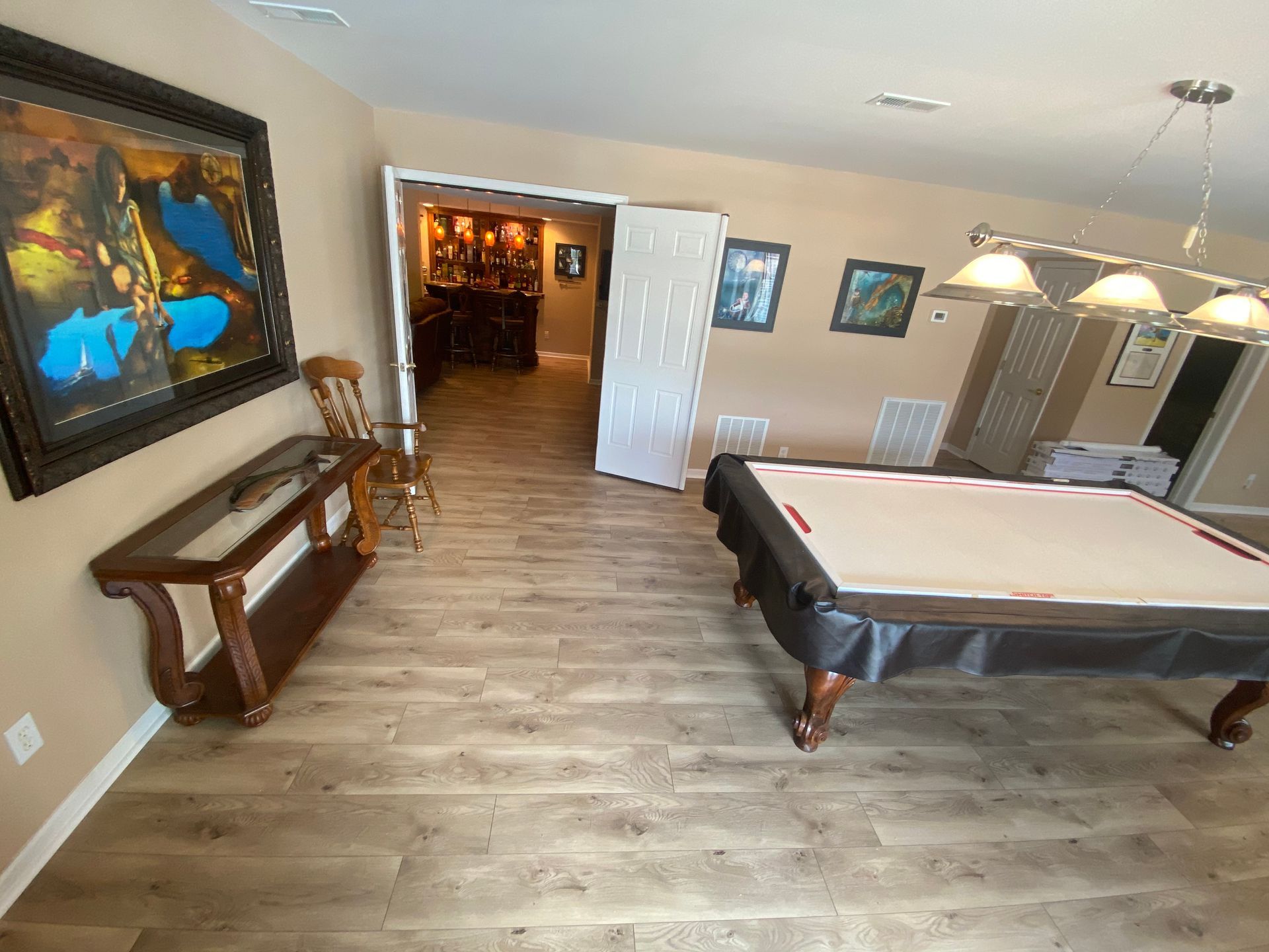 Pool table in a game room with wood floors, wall art, and a doorway to a dimly lit hall.