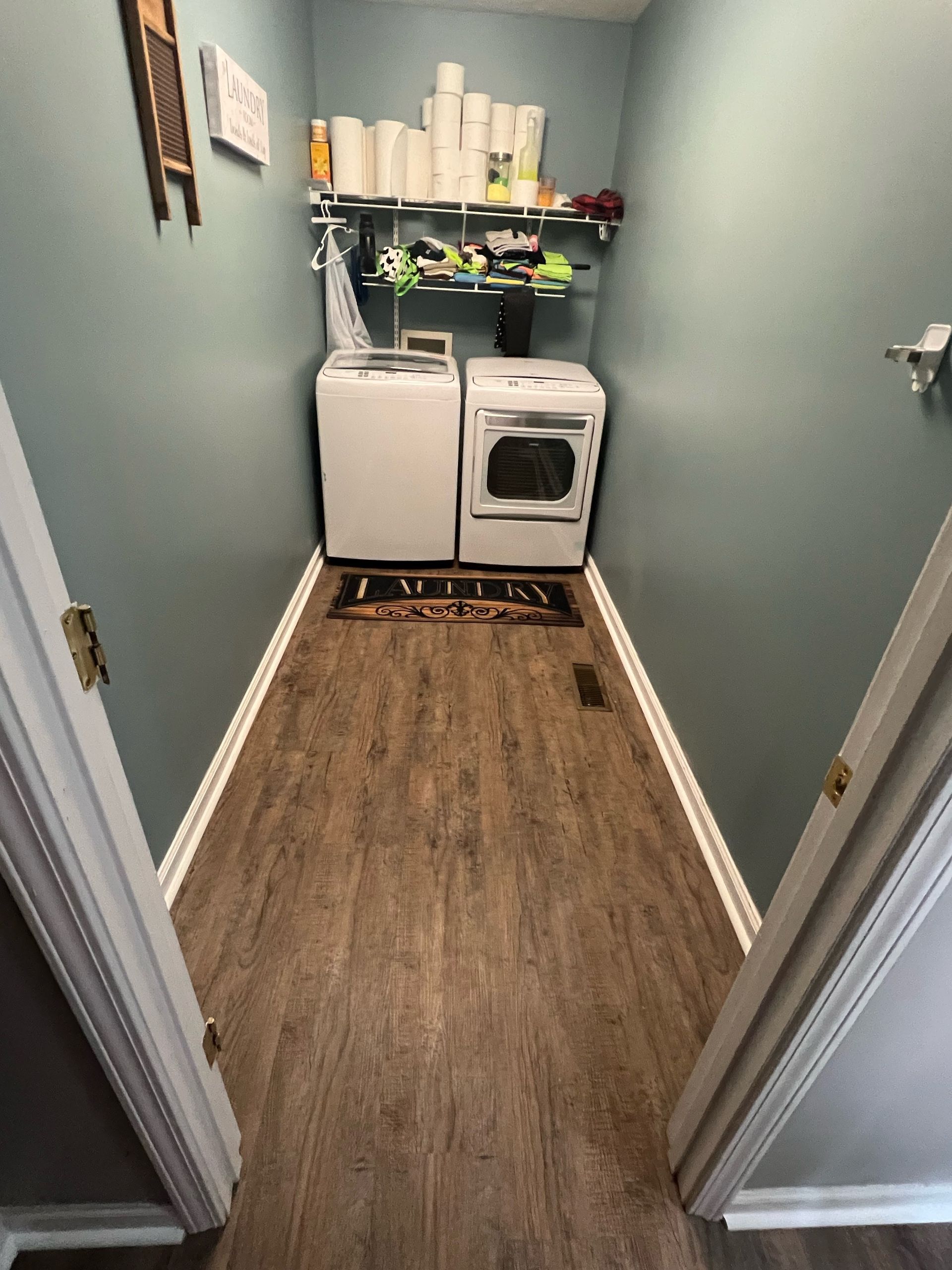 Narrow laundry room with washer, dryer, shelves, and wood-look floor.