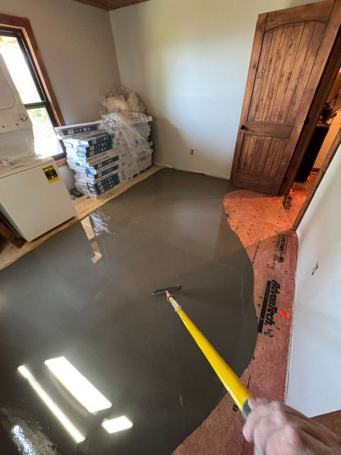 Worker smoothing wet gray floor with a squeegee in a small room with stacked materials.
