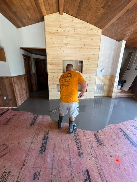 Worker in orange shirt sanding a wooden fireplace wall in a room under renovation.