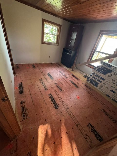 Empty room under renovation with plywood subfloor, two windows, and a wood-paneled ceiling.