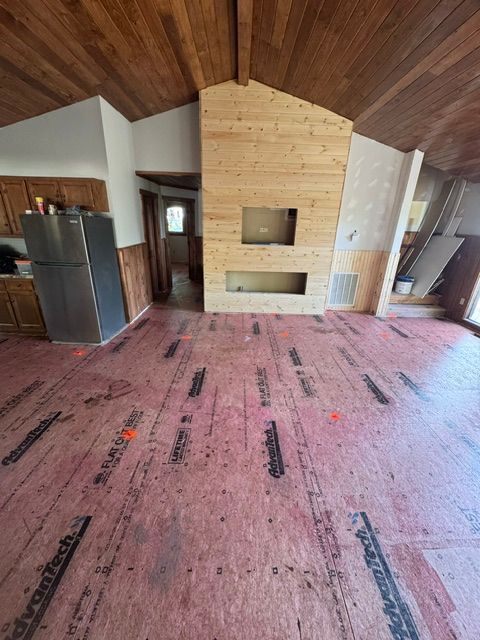 Empty living room with wood-paneled vaulted ceiling and a central stone fireplace, pink carpet, and dark refrigerator left.