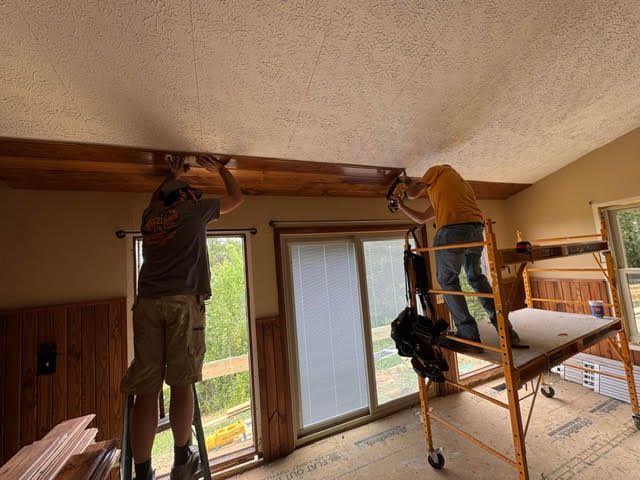 Two workers installing a wooden beam near the ceiling in a living room with a ladder and sliding glass door