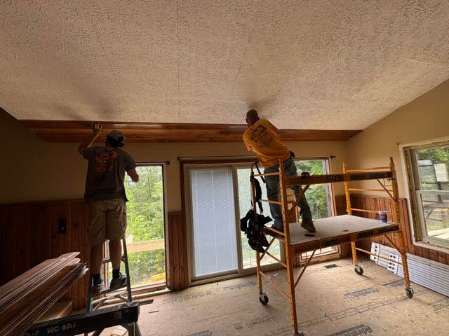 Two workers on scaffolding repainting a living room ceiling near large windows