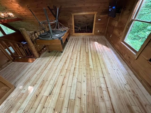 Empty wooden attic room with pine floor, exposed beams, and windows overlooking trees