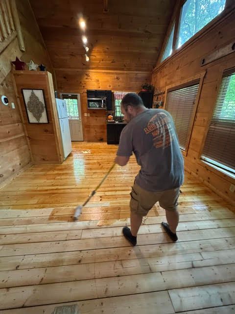 Man mopping a wooden cabin floor in a sunlit room with log walls and windows