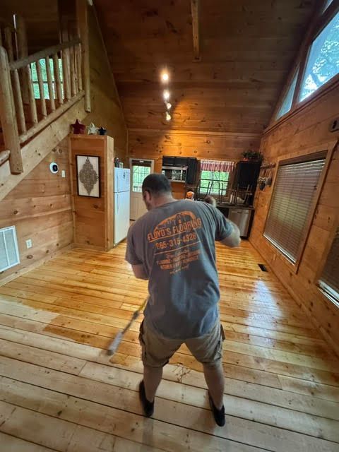 Person mopping a wooden cabin floor in a rustic room with log walls and large windows