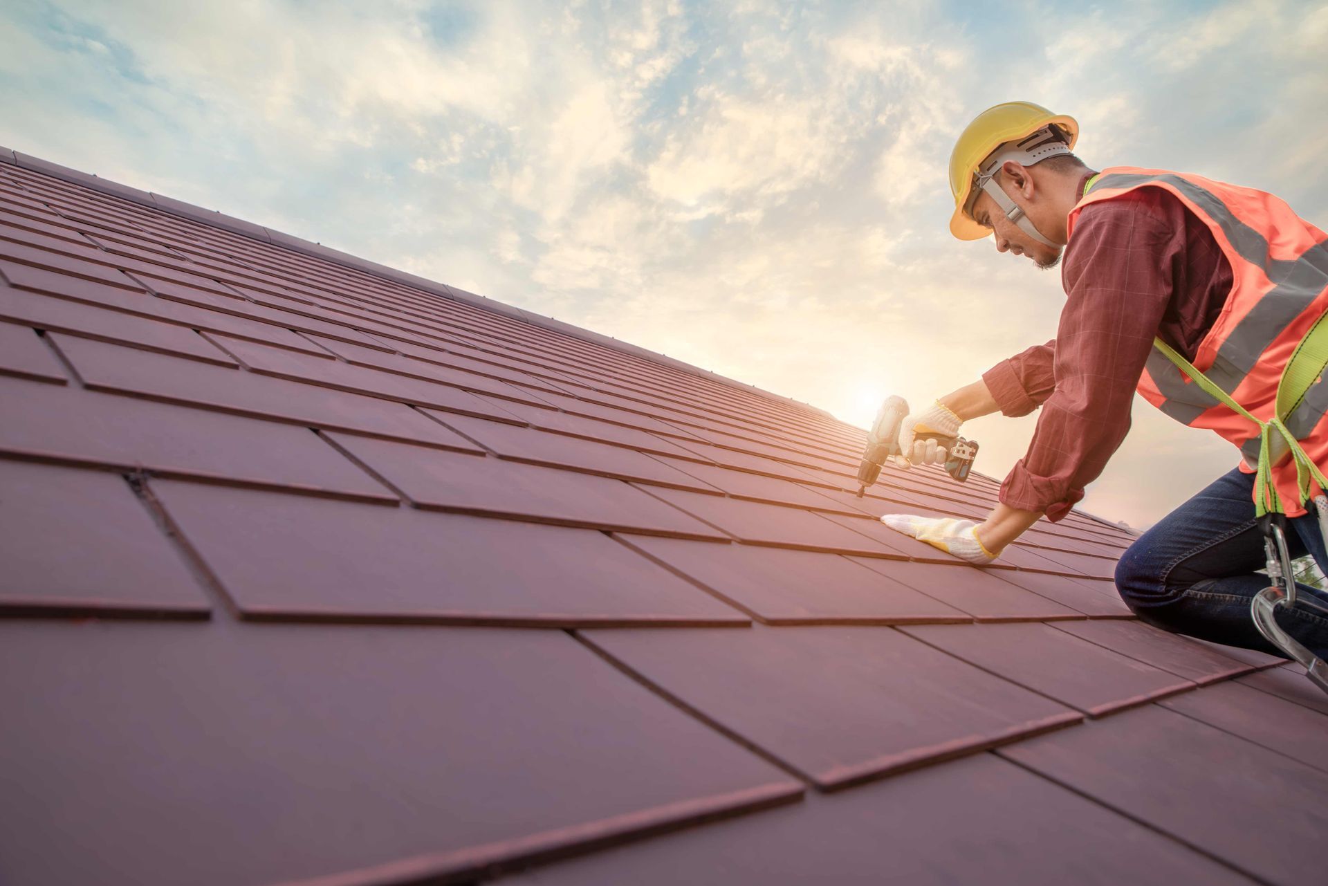Roof worker fastening red metal roofing panels on a sloped house roof with safety gear at sunrise.
