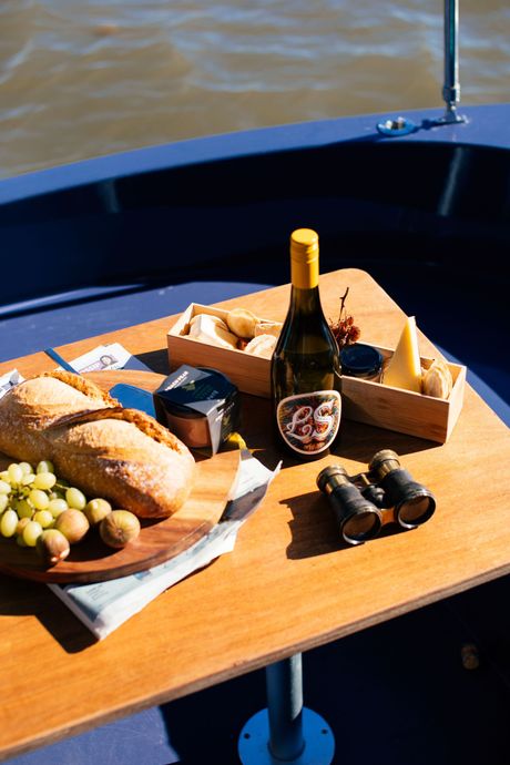 Friends dining outdoors by the water at a white table with drinks, bread, and fruit platter