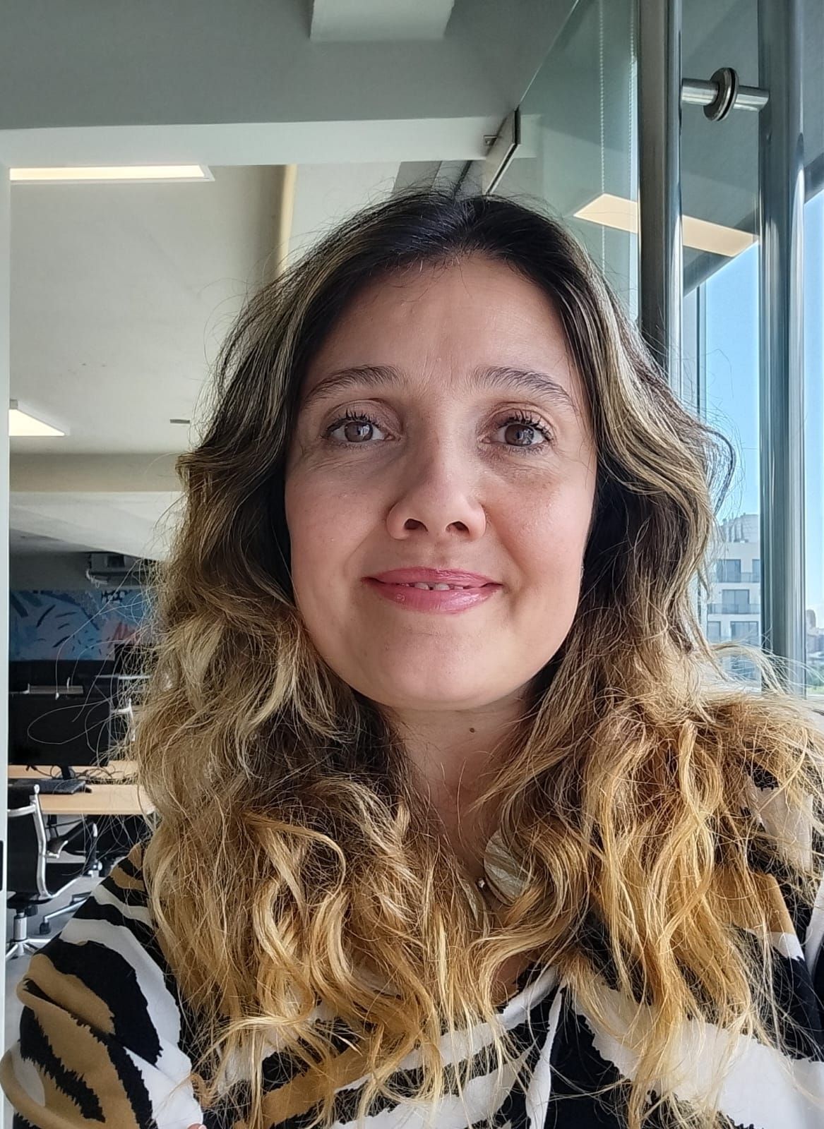 Una mujer de cabello largo y rizado, rubio y castaño, sonríe. Está en el interior, junto a una ventana