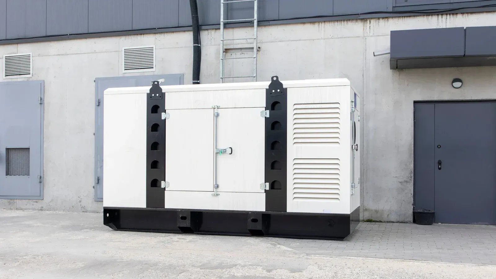 White industrial generator against a concrete building. Black support frame, exterior ventilation, ladder.