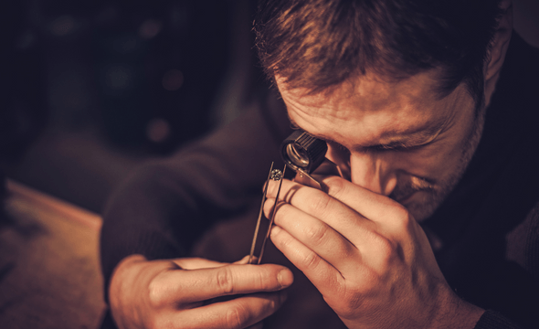 Person closely examining a small object with a magnifying glass in a dim, warm-toned setting