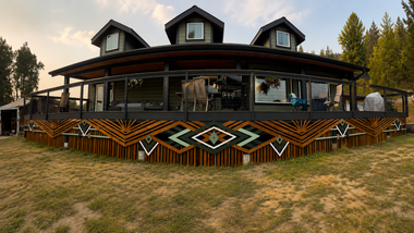 Rustic lodge with wraparound porch and geometric wood railing, set in a grassy mountain clearing at sunset