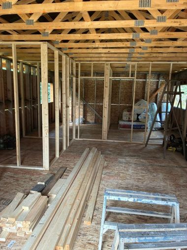 Unfinished house interior with exposed wooden framing, stacked lumber, and construction materials on the floor.