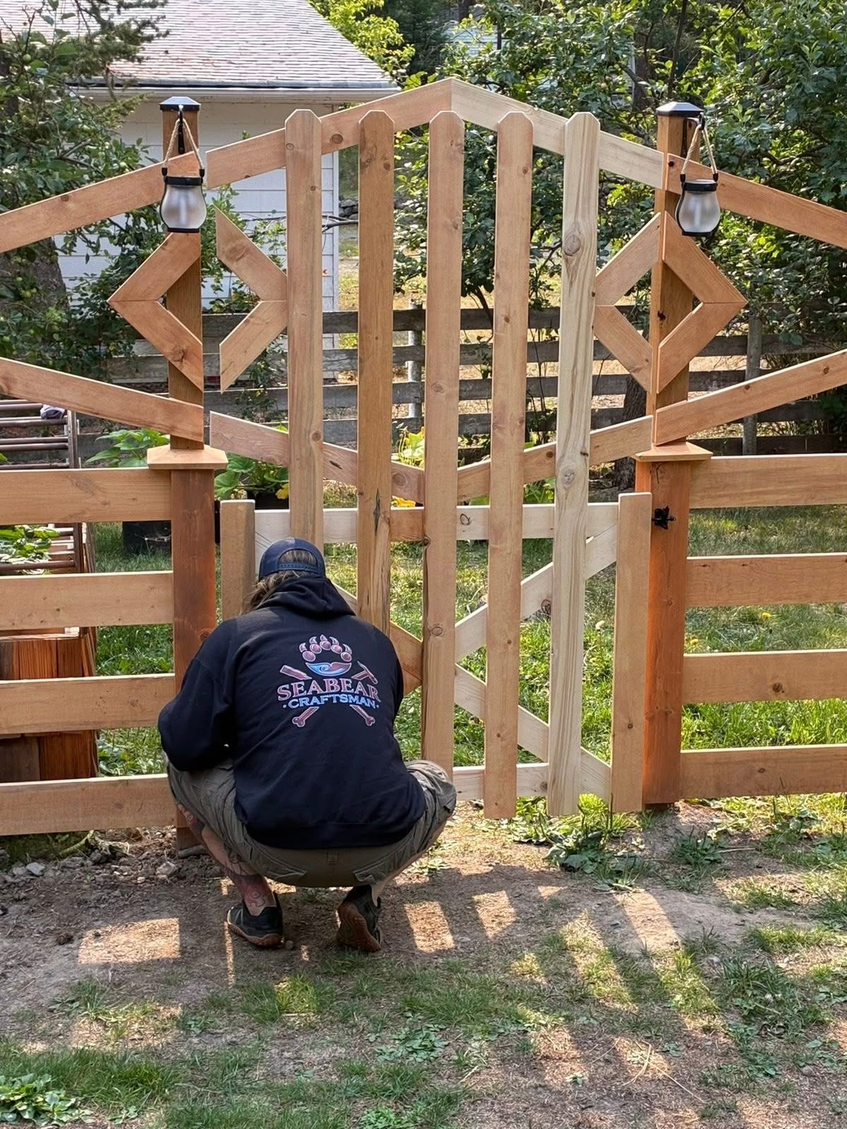 Person crouching to work on a wooden gate in a garden, with a house and trees in the background