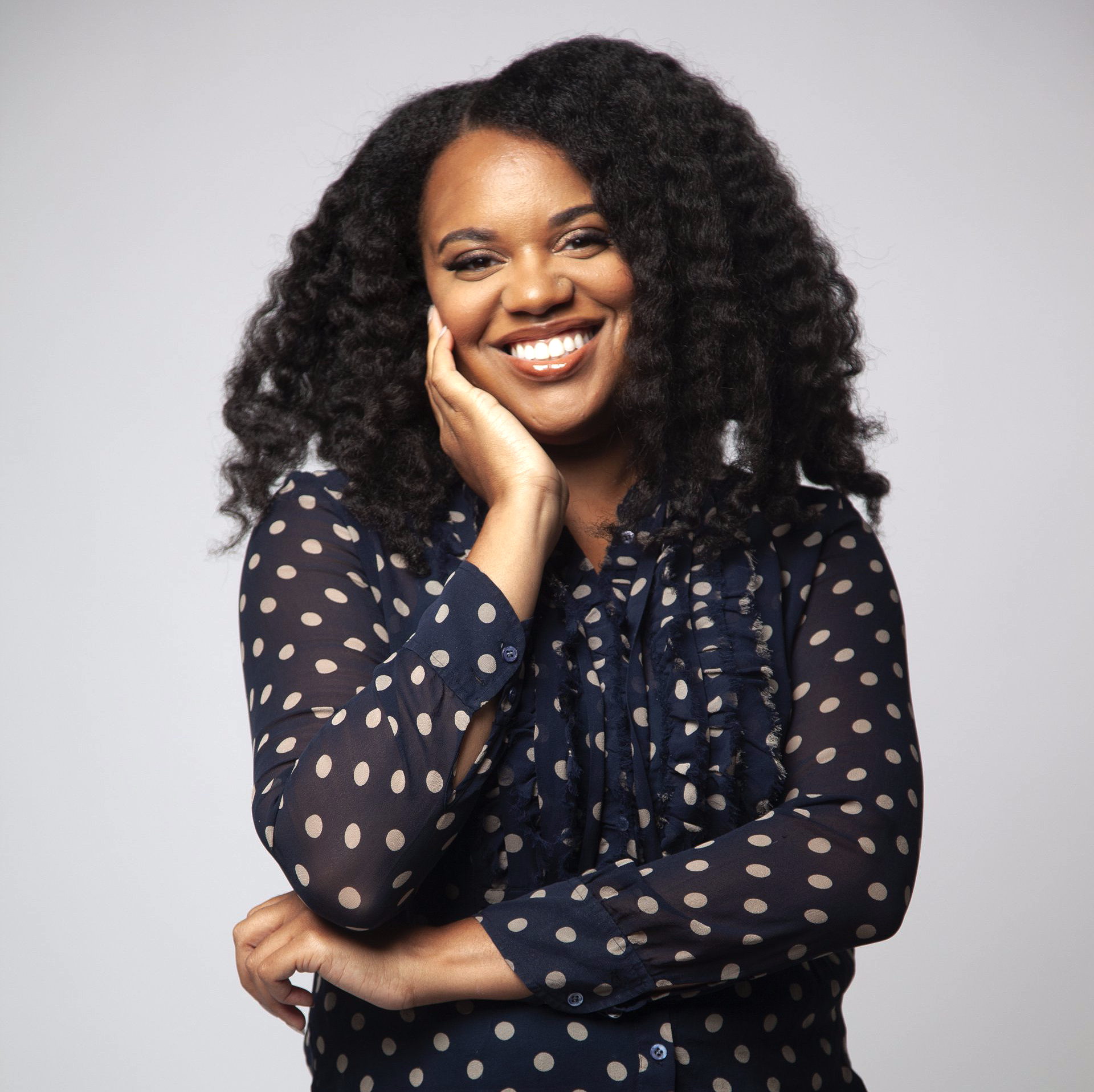 A woman with curly hair is wearing a blue polka dot shirt and smiling.
