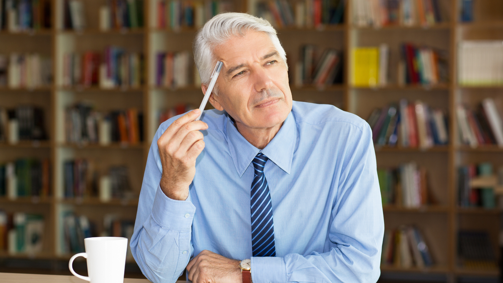 A man in a blue shirt and tie is holding a pen to his forehead and thinking