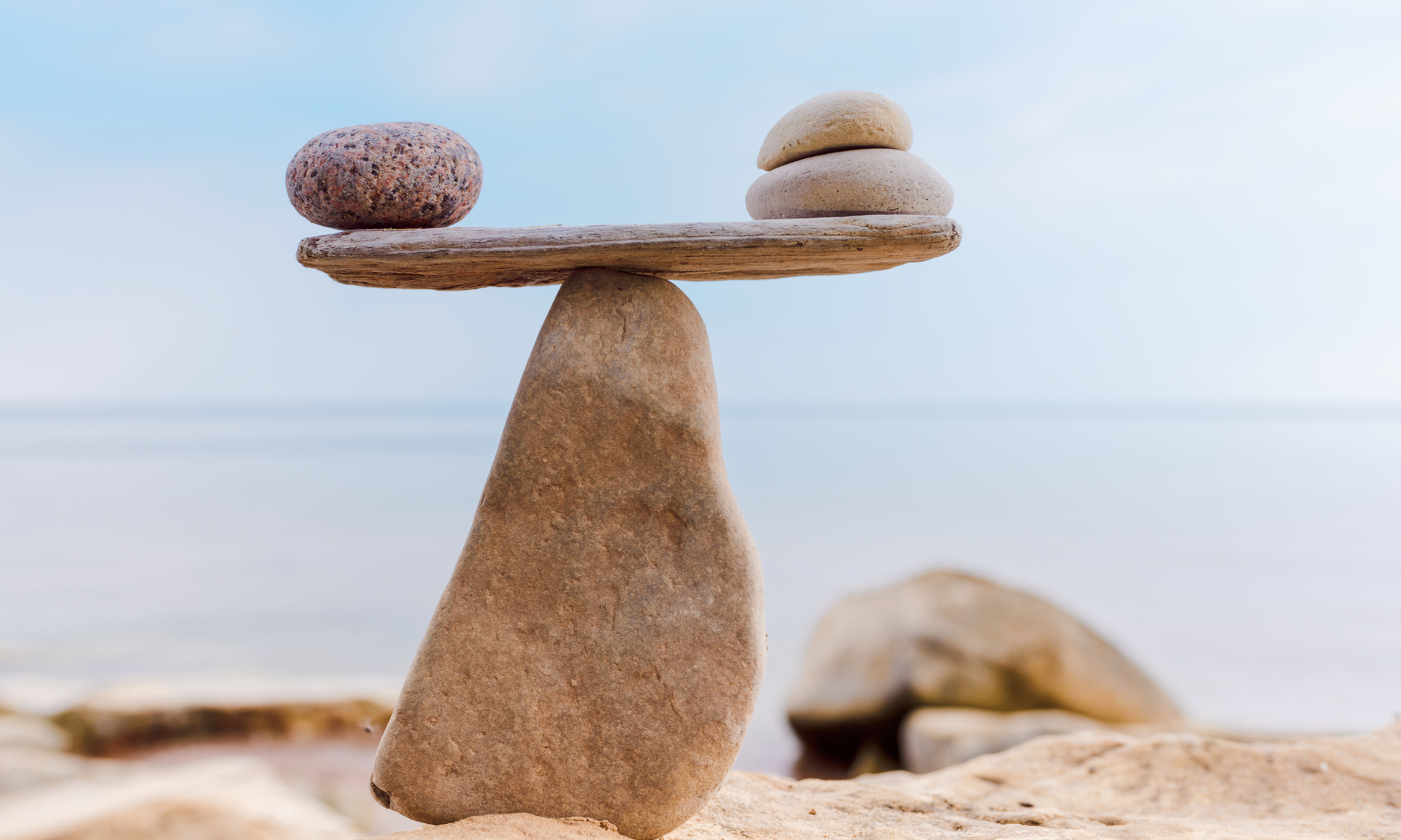 Balanced stones stacked on a beach, forming a seesaw-like shape against a calm sea background