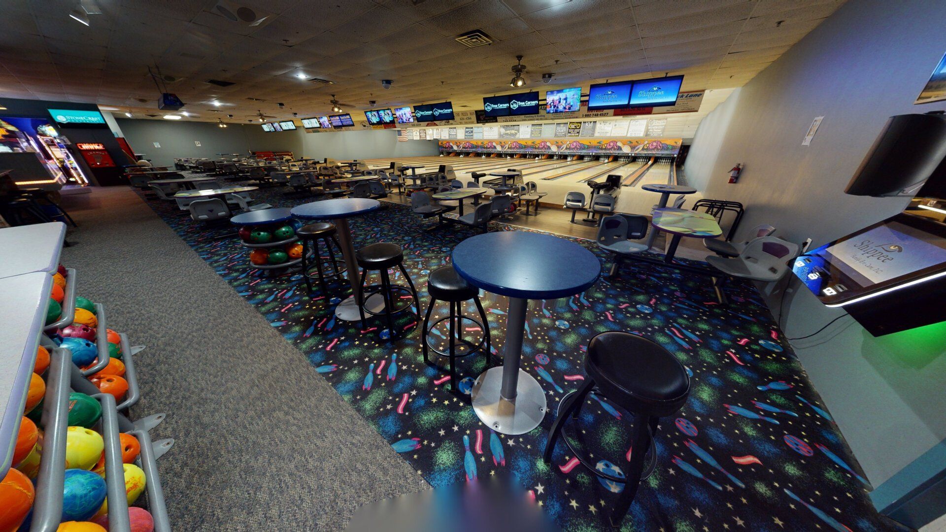 Bowling alley interior, tables, chairs, bowling balls, lanes in background, overhead screens.