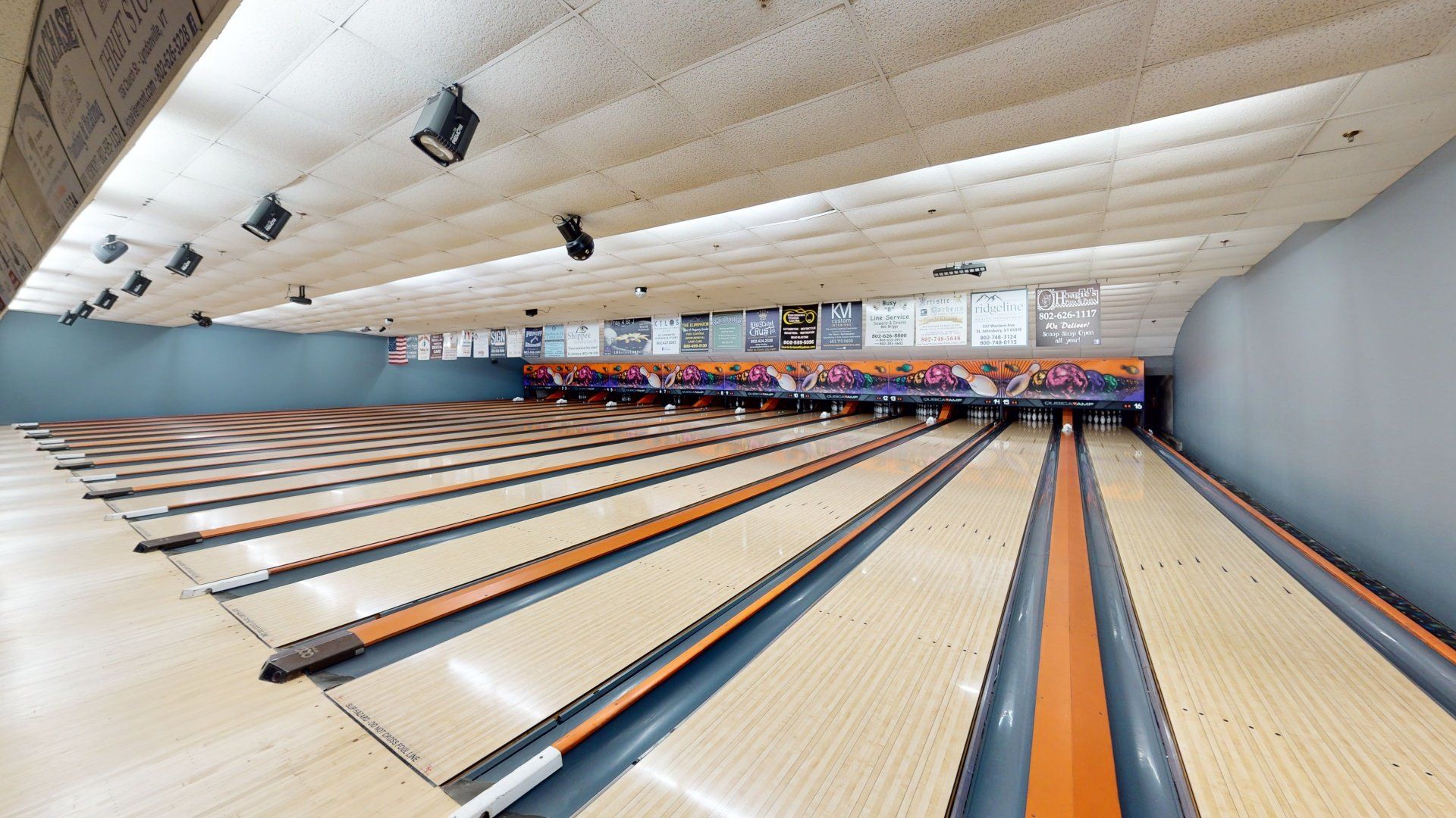 Bowling alley with multiple lanes. Tan lanes with orange dividers, gray walls, and light fixtures.