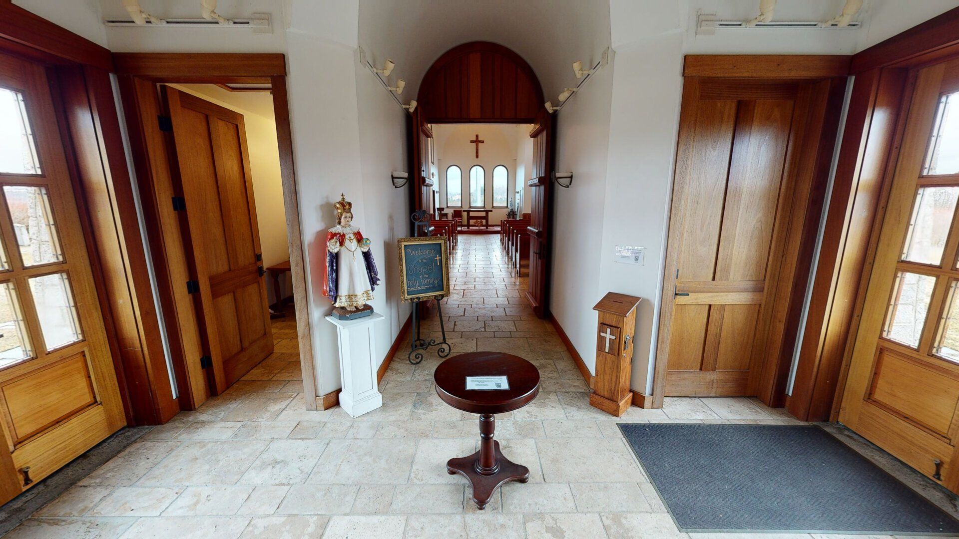 Interior hallway leading to a church sanctuary. Two wooden doors flank the entrance, a statue, table.