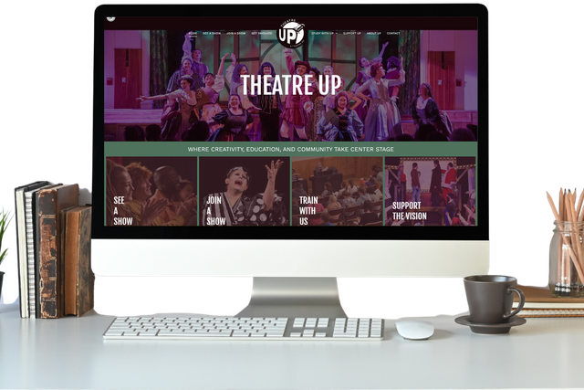 Desktop monitor displaying a theater website on a white desk with books, coffee cup, and stationery