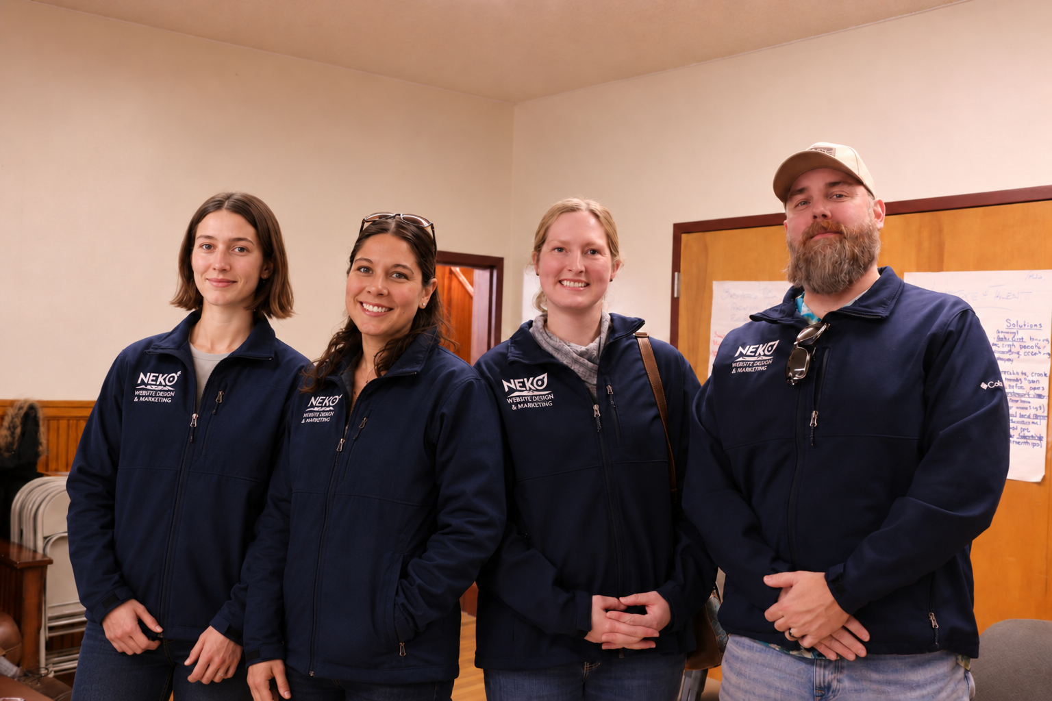 Four people in navy jackets are standing indoors, smiling in a small room.