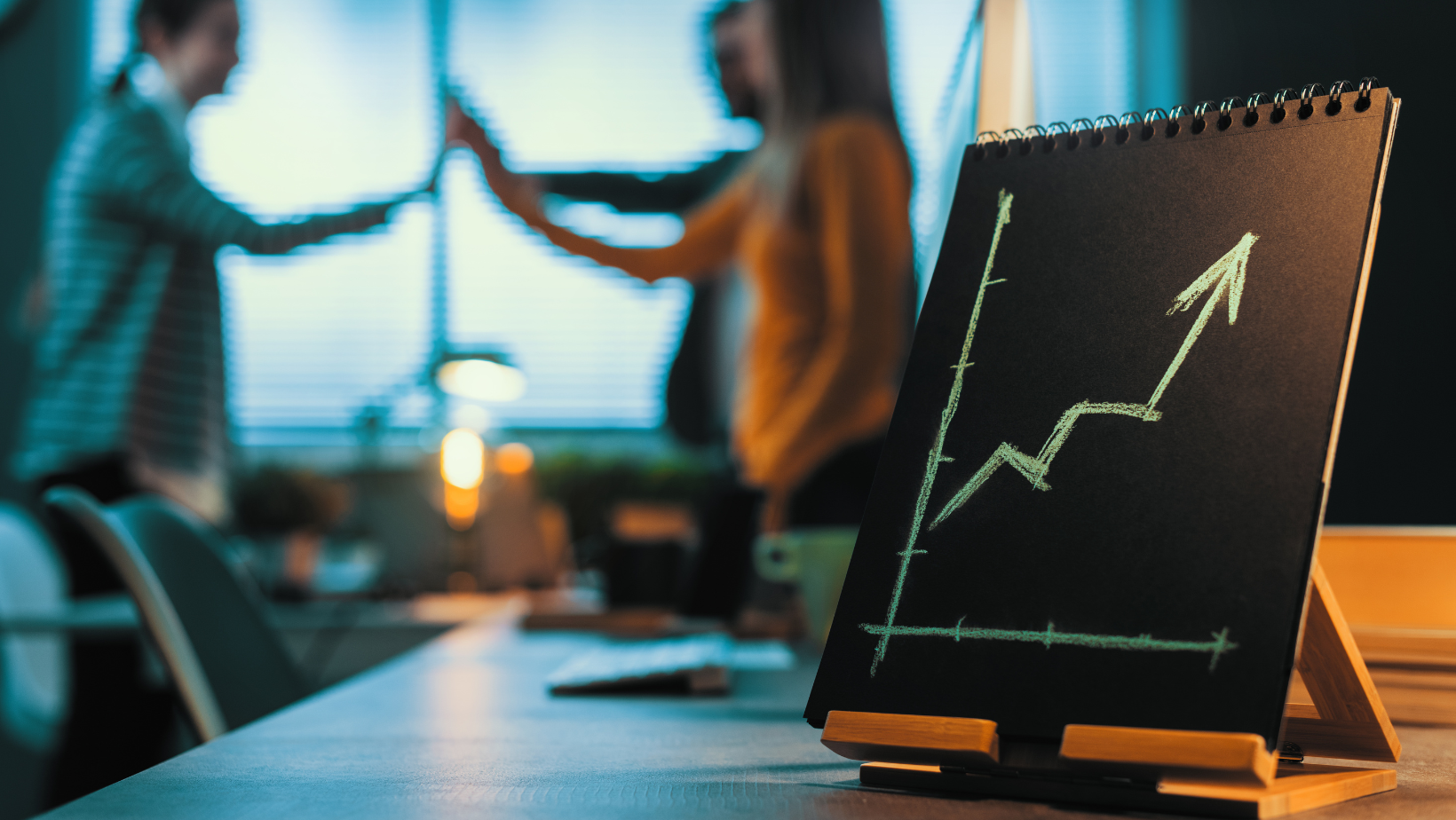 A notebook with a growth chart on it is sitting on a desk. People in the background are celebrating.