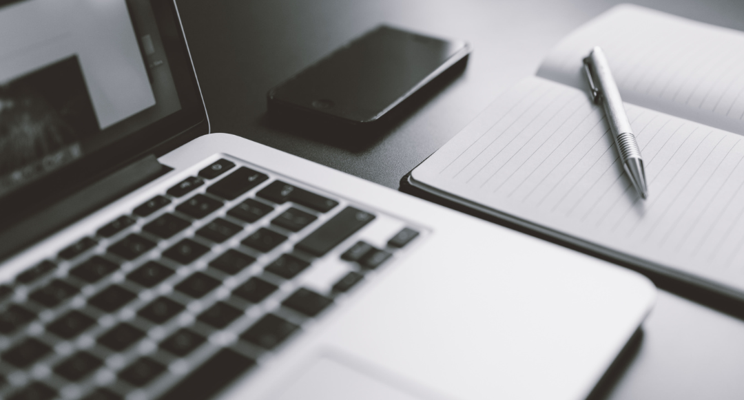 A laptop, cell phone and pen and paper sitting on a desk.
