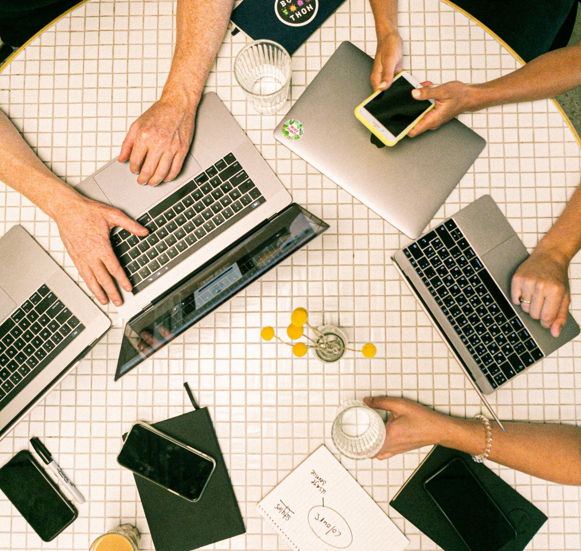 An overhead shot of a table with many hands using laptops, tablets and phones for digital marketing.