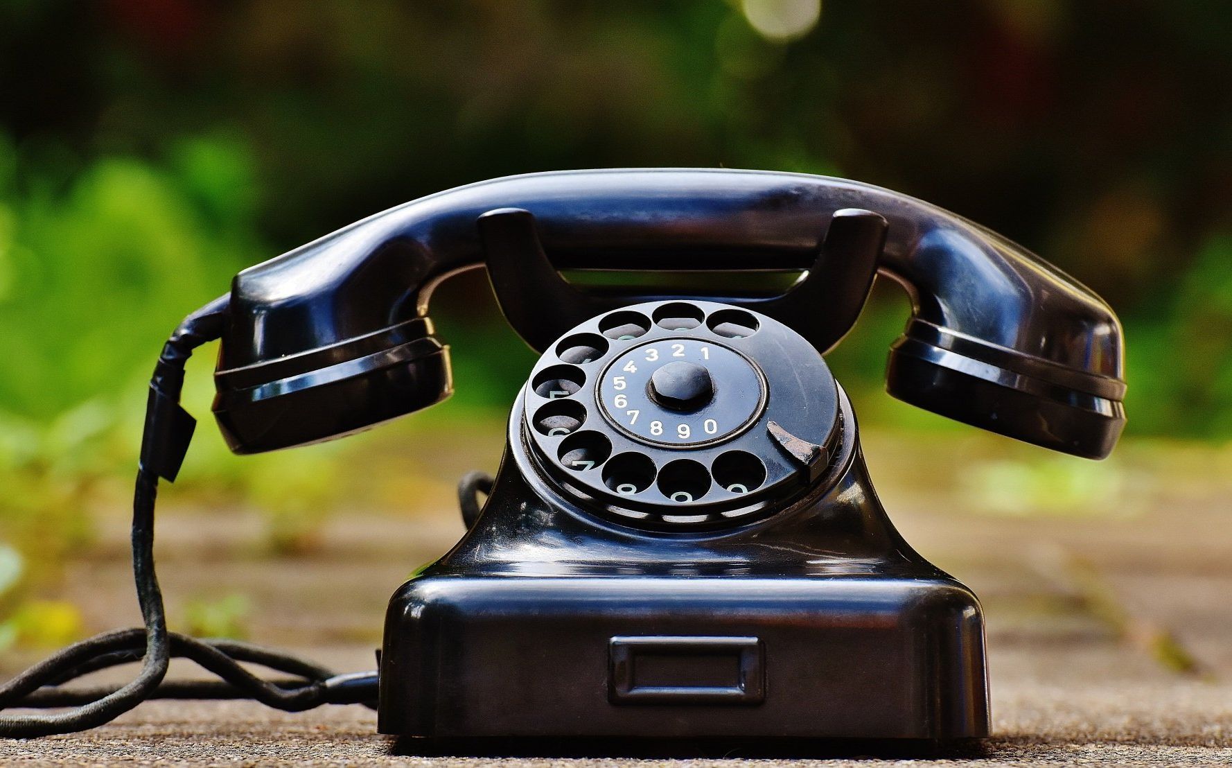 Black rotary phone with receiver off-hook, sitting on a wooden surface outdoors with blurred green background.