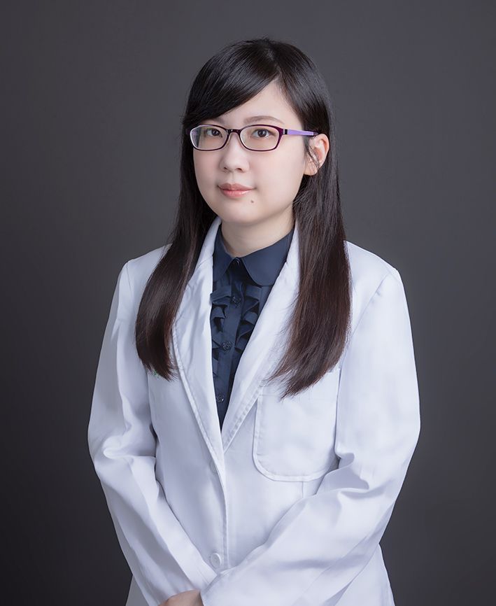 A woman in a white coat and glasses is standing in front of a black background.