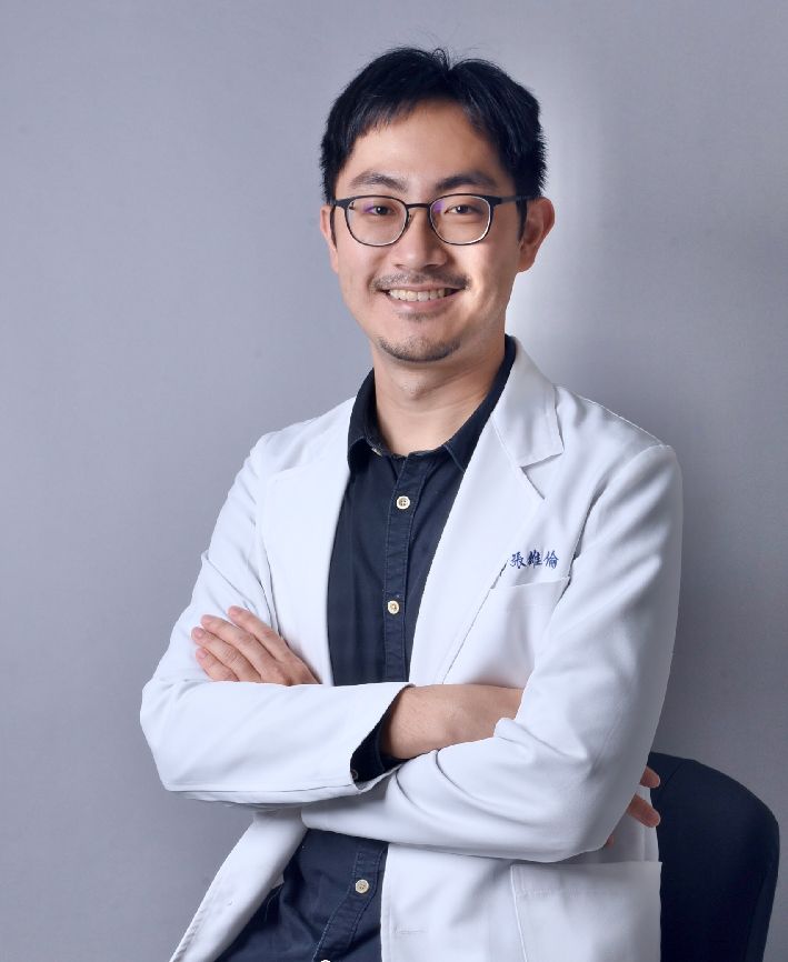 A man in a white coat and glasses is sitting in a chair with his arms crossed.