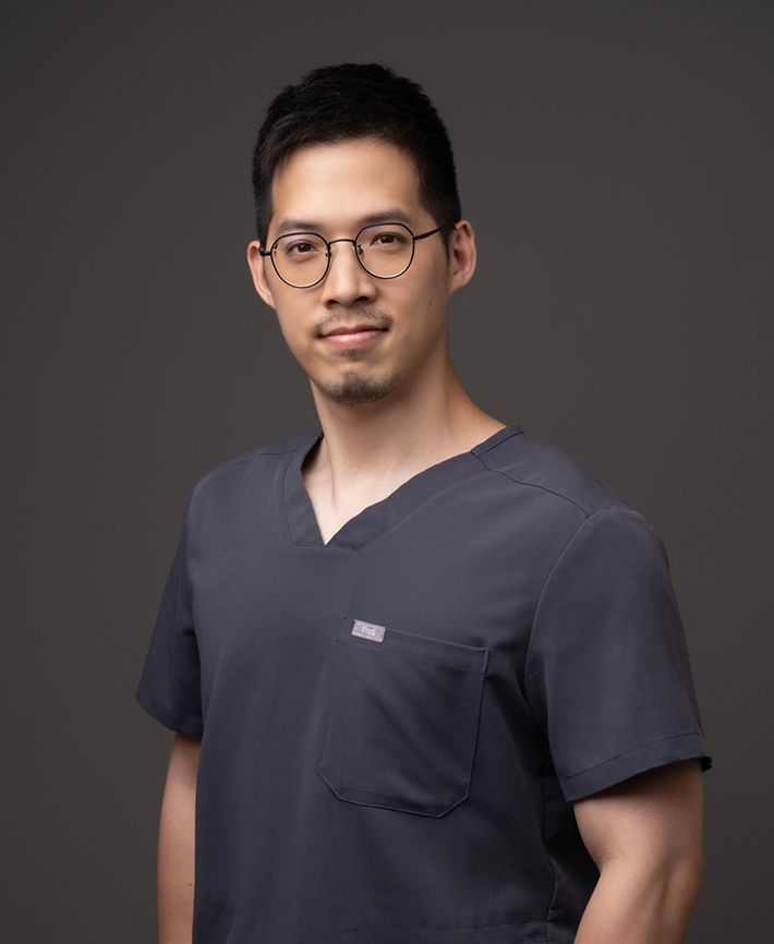 A man wearing glasses and a scrub top is standing in front of a gray background.