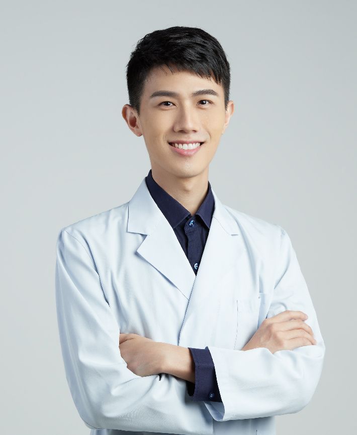 A young man in a lab coat and tie is standing with his arms crossed.