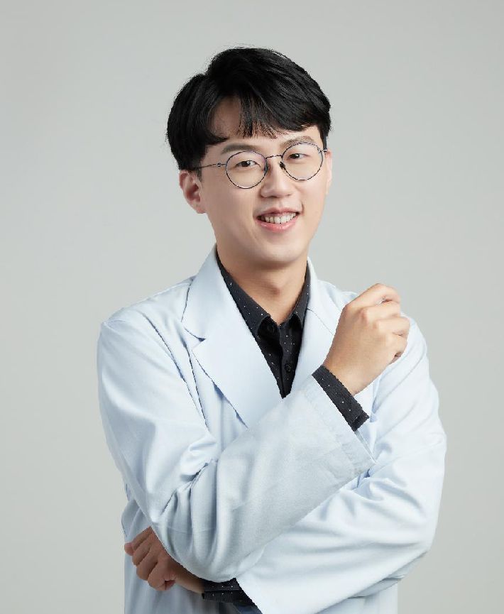 A young man wearing glasses and a white coat is standing with his arms crossed.