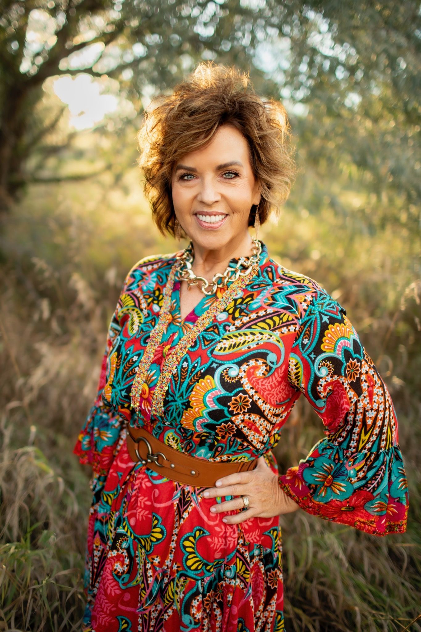 Woman wearing colorful patterned dress outdoors, smiling, with hand on hip.