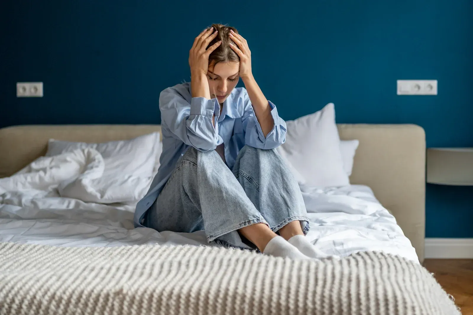 Woman sitting on a bed, holding her head. She looks distressed in a bedroom with blue walls.