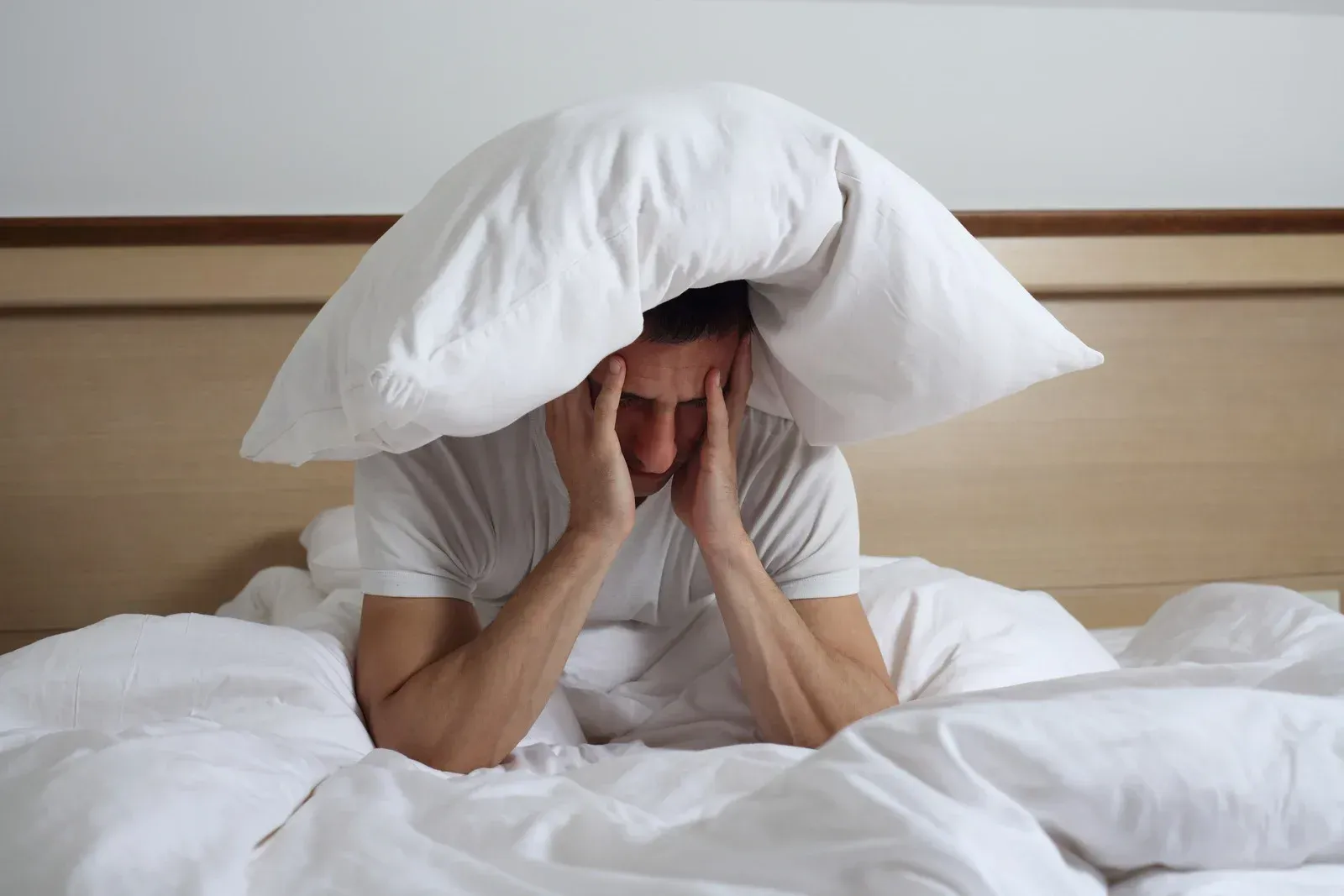 Man in bed with pillow over head, appears stressed and holding temples.