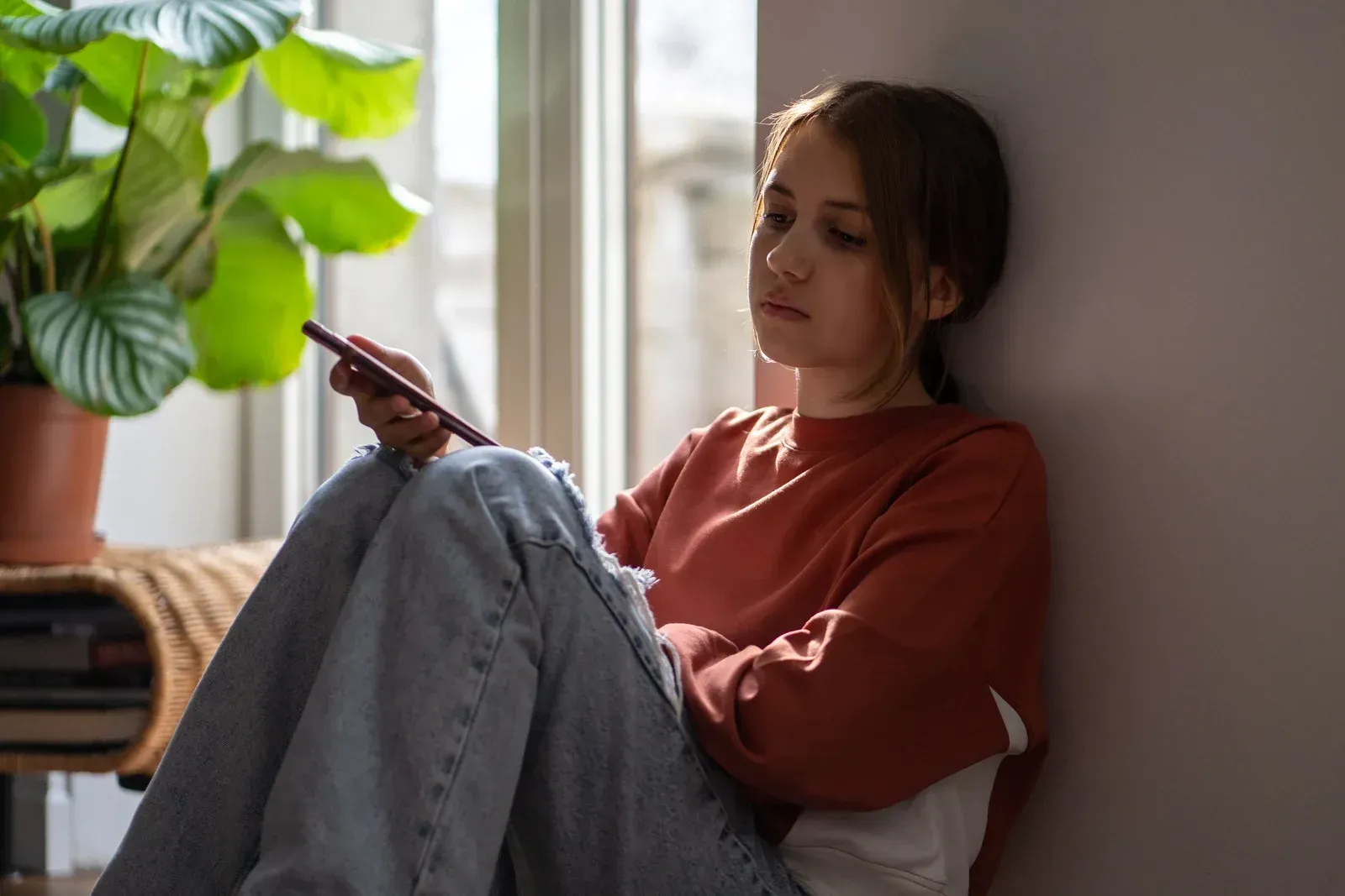Teen girl sits by a window, looking at her phone, looking sad.