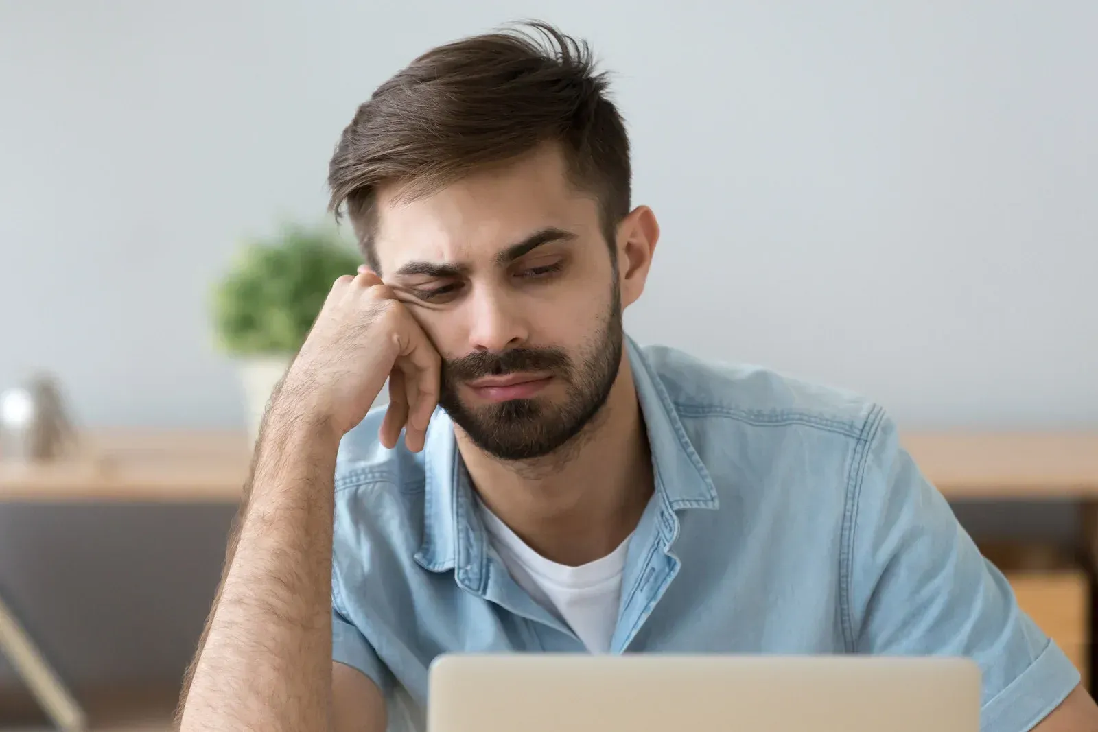 Man with beard resting his head on hand, looking tired in front of a laptop.