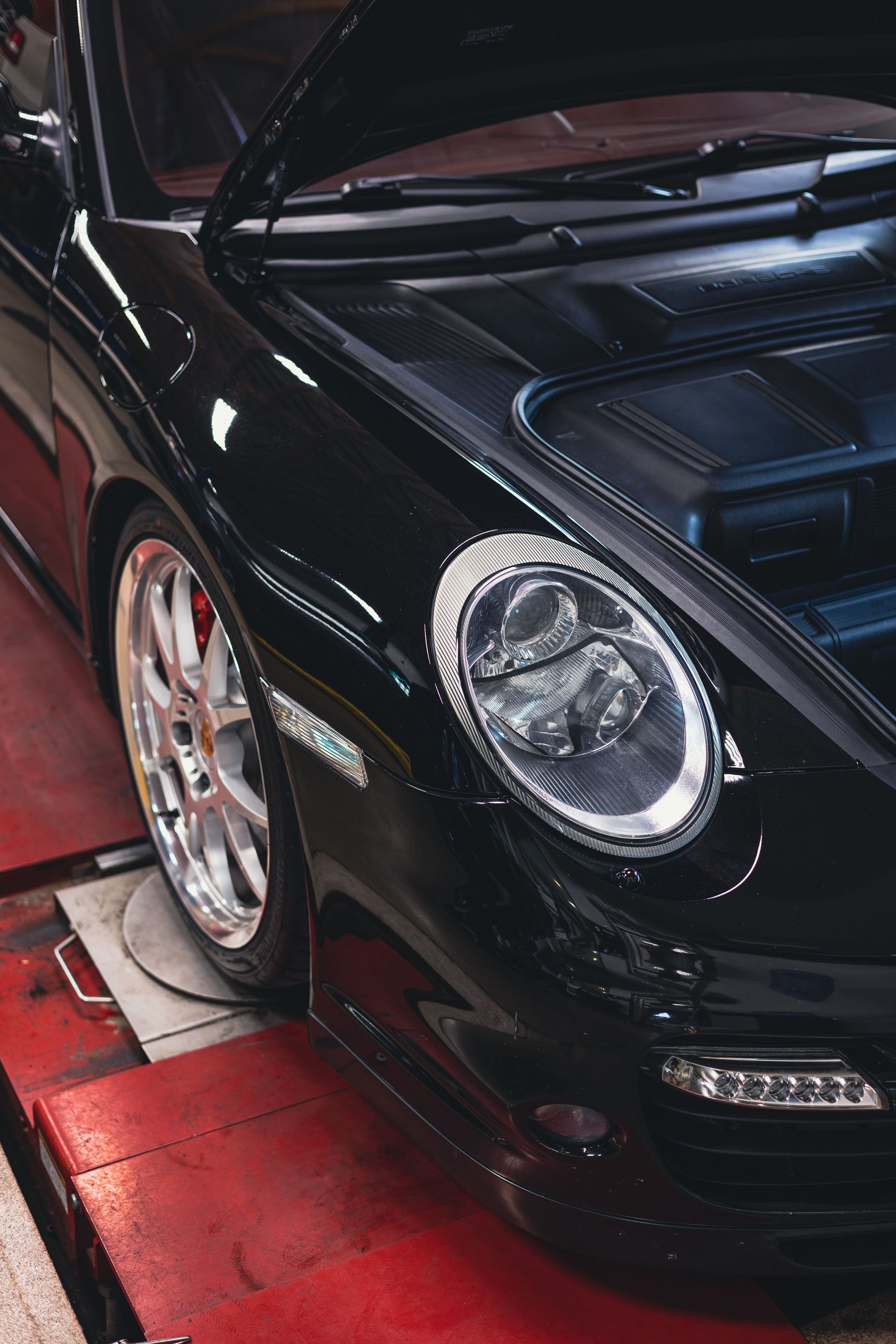 Black sports car front end with headlight detail, parked indoors on a red floor.