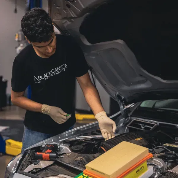 Mechanic working under open car hood in a garage, wearing gloves and a black T-shirt.