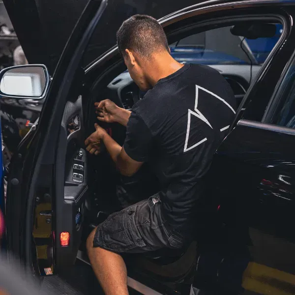 Person sitting in a black car, reaching toward the steering wheel through the open door.