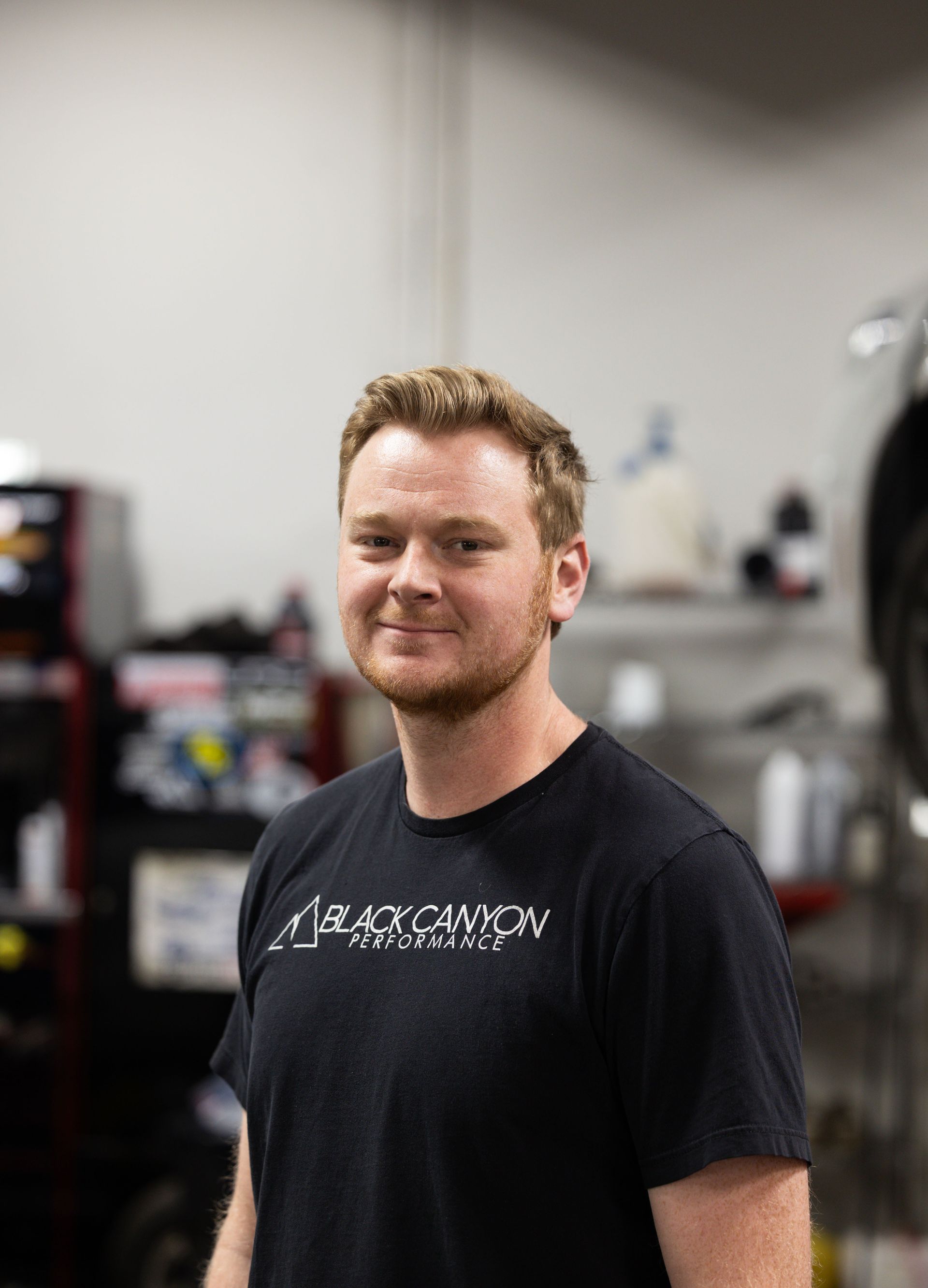 Smiling person in a black T-shirt standing in a gym, with equipment blurred in the background.