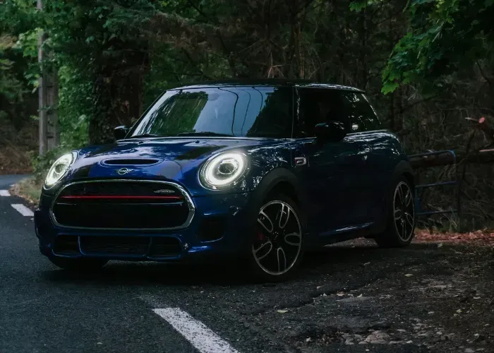 Blue Mini Cooper parked on a forest road, headlights on, with a white lane line in front.