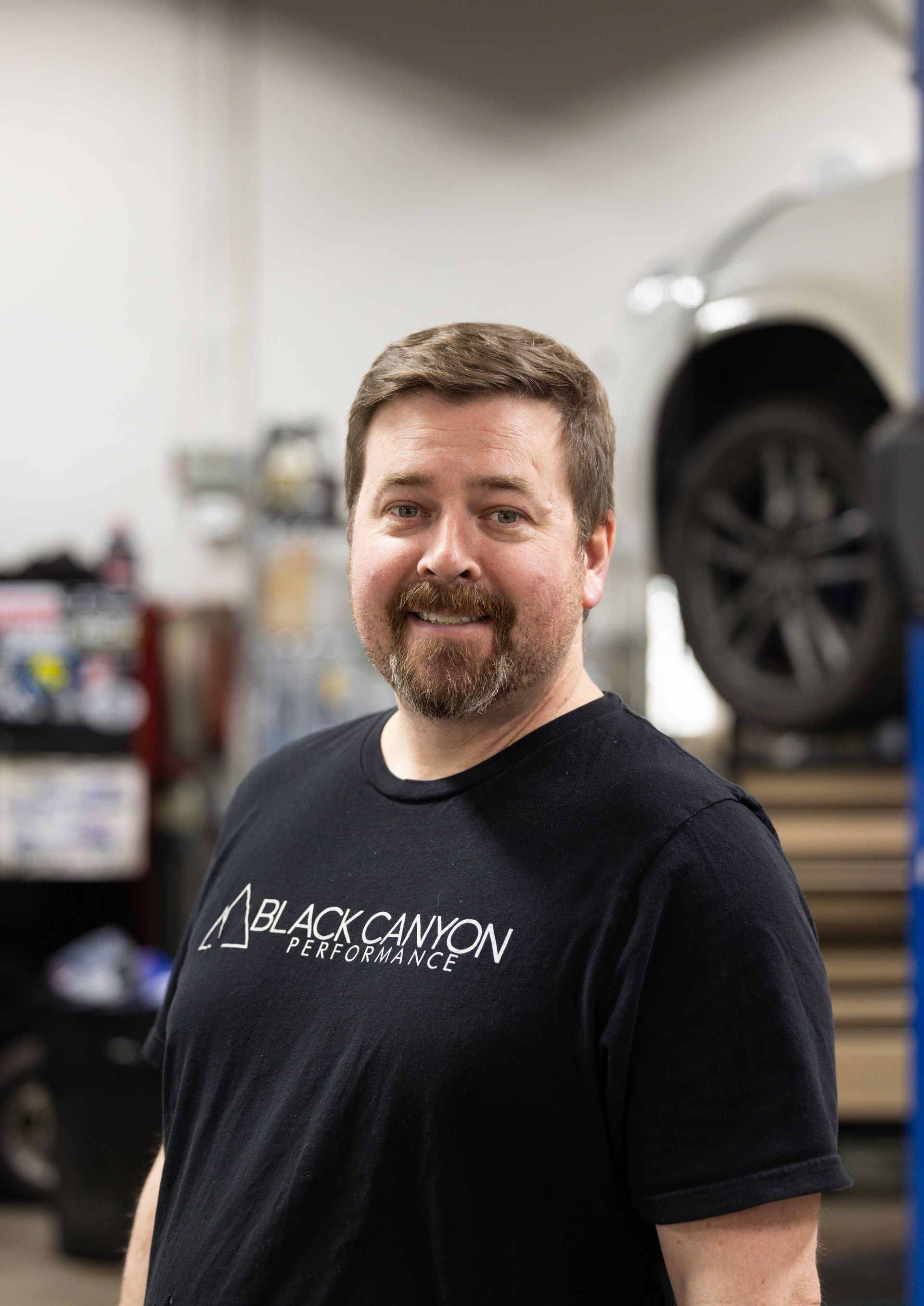 Man standing in a garage, smiling in a black T-shirt beside a vehicle lift