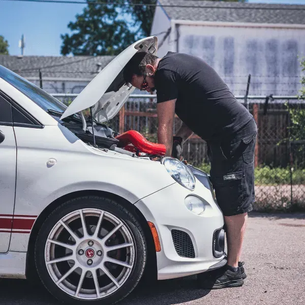 Man in black shirt working under the hood of a white sports car on a sunny street